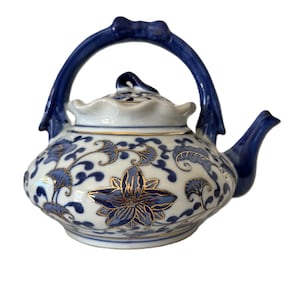 Könnte beinhalten: Eine weiß-blaue Keramik-Teekanne mit gebogenem Henkel und Ausguss. Die Teekanne ist mit blauen Blumenmustern und goldenen Akzenten verziert. Der Deckel hat einen dekorativen Knauf. Ein klassisches Stück für Teeliebhaber.