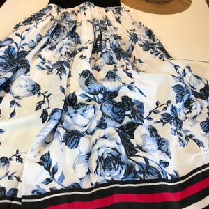 May include: A black top with a white and blue floral print maxi skirt. The skirt has a pattern of blue roses on a white background. A straw hat is next to the dress. The dress is suitable for summer.
