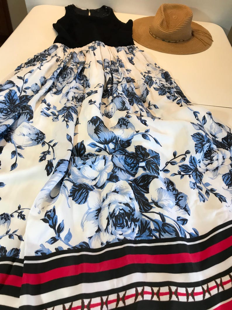 May include: A black top with a white and blue floral print maxi skirt. The skirt has a pattern of blue roses on a white background. A straw hat is next to the dress. The dress is suitable for summer.