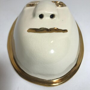 May include: White ceramic butter dish with a gold rim and a face design. The face has two small holes for nostrils, a gold line for a mouth, and gold accents for eyes.
