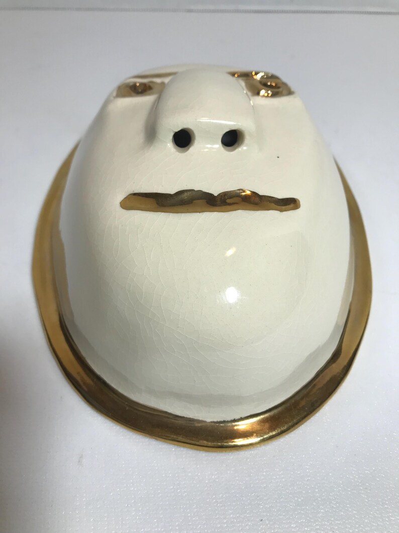 May include: White ceramic butter dish with a gold rim and a face design. The face has two small holes for nostrils, a gold line for a mouth, and gold accents for eyes.