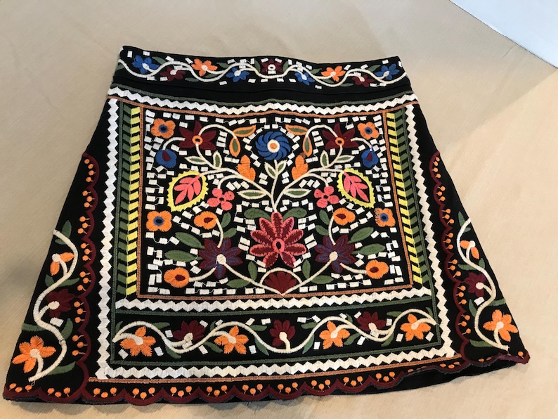 Puede incluir: Una minifalda negra con coloridos patrones florales bordados. La falda tiene un dobladillo festoneado y una cintura negra.
