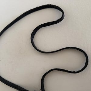 Puede incluir: Primer plano de un cord&oacute;n negro, fino y flexible, posiblemente de cuero o material similar, sobre un fondo blanco. El cord&oacute;n est&aacute; curvado y en bucle, con una superficie texturizada. La imagen destaca los detalles del cord&oacute;n.