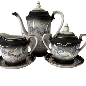 Puede incluir: Juego de té de porcelana vintage con tetera, jarra de crema y azucarero con tapa. El juego presenta un diseño degradado en blanco y negro con motivos de dragones en tonos grises y azules, acentuado con ribetes dorados.
