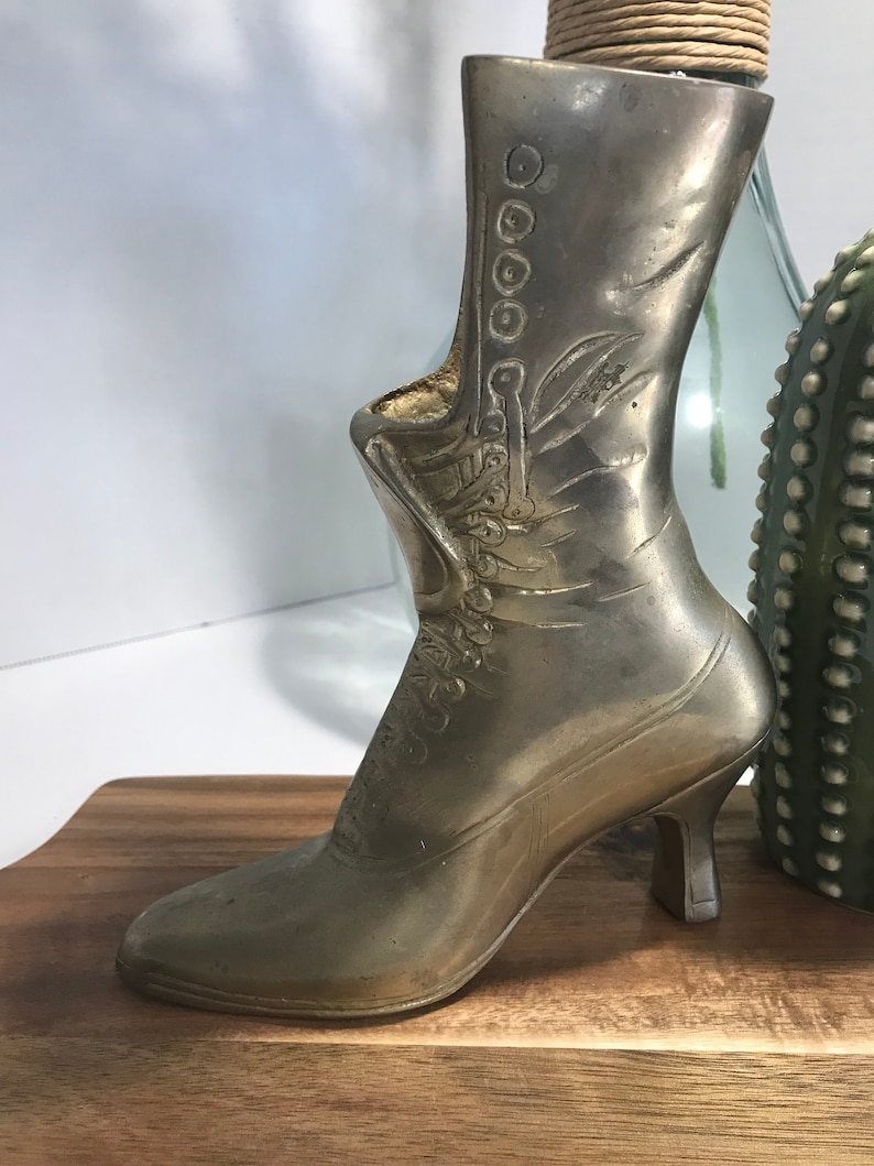 May include: A silver metal boot shaped vase with a high heel. The boot has a decorative design on the front and a row of buttons on the side.