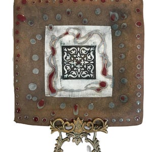 May include: A square ceramic plate with a brown background and a white square in the centre. The central square features a black and white ornate design. The plate is supported by a decorative metal stand.