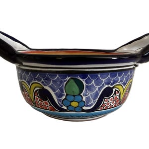 May include: A hand-painted ceramic bowl with two handles. The bowl features a blue background with a scale pattern and colorful floral and geometric designs. The handles are black, and the rim has a dark blue border.
