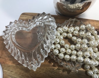 Può includere: Due piatti di vetro trasparente a forma di cuore con una collana di perle bianche su una superficie di legno.