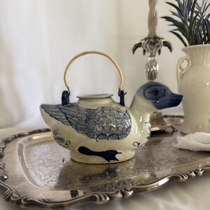 May include: A ceramic teapot shaped like a duck with a cream-coloured body and blue painted details including feathers, a beak, and feet. The teapot features a circular lid and a thin, arched, light-coloured handle attached to two blue loops.