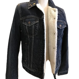 May include: Dark blue denim jacket with a white sherpa lining. The jacket has two chest pockets, two side pockets, and gold star embellishments on the collar. The jacket is displayed on a mannequin.