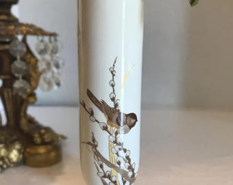 Puede incluir: Un jarrón cilíndrico alto de cerámica color crema con una delicada ilustración de un pájaro marrón posado sobre ramas de sauce. El diseño incluye sutiles detalles dorados y un acabado suave y brillante, con un borde acampanado y base estable.