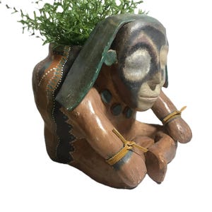 May include: A brown clay planter shaped like a seated figure with a painted face, dark green head covering, and tan leather ties on the arms. The figure features black and white facial markings and holds a small green plant in the top opening.
