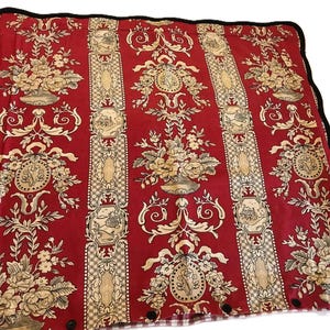 May include: Red fabric with a floral and ornamental design. The fabric features cream-colored floral arrangements, medallions, and vertical panels. A black scalloped border frames the edges. The fabric is likely used for home decor.
