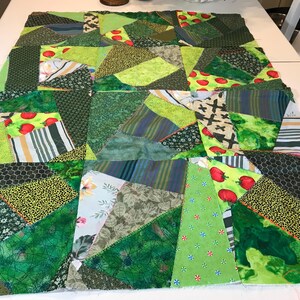 May include: A patchwork quilt in various shades of green, featuring a mix of patterns including floral, stripes, and geometric shapes. Some patches have red tomato designs. The quilt is laid out on a white surface.