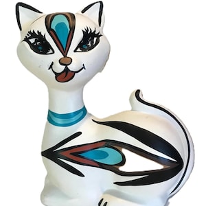 Może przedstawiać: Biała ceramiczna figurka kota z czarnymi, niebieskimi i brązowymi akcentami dekoracyjnymi. Kot ma duże oczy, niebieski kołnierz i figlarny wyraz twarzy. Projekt zawiera stylizowane wzory przypominające pióra.