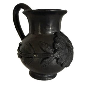 May include: A dark grey ceramic jug with a handle and a wide mouth. The jug features a floral design on the front and a decorative band around the middle. The jug is likely used for serving beverages.