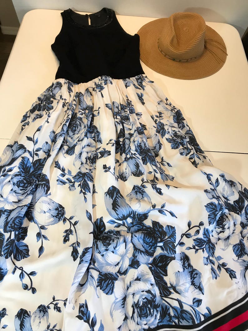 May include: A black sleeveless dress with a full skirt, featuring a white background and blue floral print. A straw sun hat with a wide brim is next to the dress. The dress has a beaded neckline.