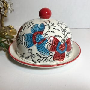 May include: A ceramic butter dish with a domed lid and matching plate. The dish is white with a red rim and features a floral design in red and blue. A red knob tops the lid.