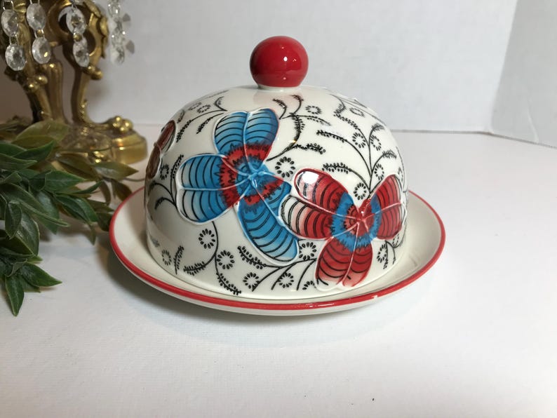 May include: A ceramic butter dish with a domed lid and matching plate. The dish is white with a red rim and features a floral design in red and blue. A red knob tops the lid.