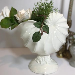 Puede incluir: Un jarrón de cerámica blanca con forma de concha de nautilo y base acampanada. El jarrón contiene rosas blancas artificiales con hojas verdes y una rama de follaje verde. La superficie del recipiente presenta nervaduras curvas en relieve.