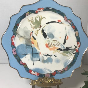 May include: A white ceramic plate with a blue rim and a gold trim. The plate features a watercolour painting of a bird with a red, yellow, and green design. The plate also has the Chinese character for "year" in black ink.