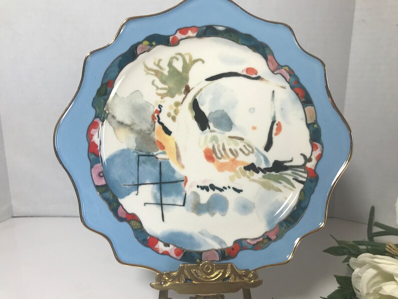 May include: A white ceramic plate with a blue rim and a gold trim. The plate features a watercolour painting of a bird with a red, yellow, and green design. The plate also has the Chinese character for "year" in black ink.