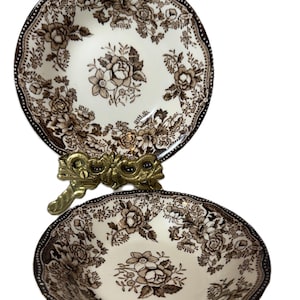 Może przedstawiać: Dwie miski ceramiczne w stylu vintage z kwiatowym wzorem w kolorze brązowym i kremowym. Miski mają falisty brzeg i ciemnobrązową obwódkę. Pomiędzy miskami znajduje się ozdobny mosiężny stojak.