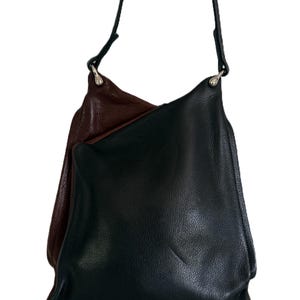 Puede incluir: Un bolso de hombro de cuero negro con una correa larga y delgada. El bolso tiene un dise&ntilde;o suave y holgado. Una segunda bolsa, de color burdeos oscuro, es parcialmente visible detr&aacute;s de la bolsa negra. Las bolsas est&aacute;n hechas de cuero texturizado.