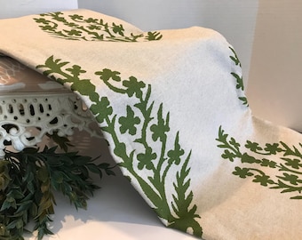 Könnte beinhalten: Ein beigefarbener Tischläufer aus Leinen mit einem grünen Blumenmuster. Das Muster zeigt stilisierte Blätter und Blüten.
