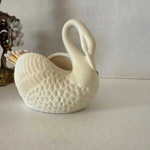 May include: Cream-colored ceramic swan figurine with textured feather detailing. The swan has a curved neck and a small opening on its back. A decorative brass and crystal candle holder is partially visible in the background.