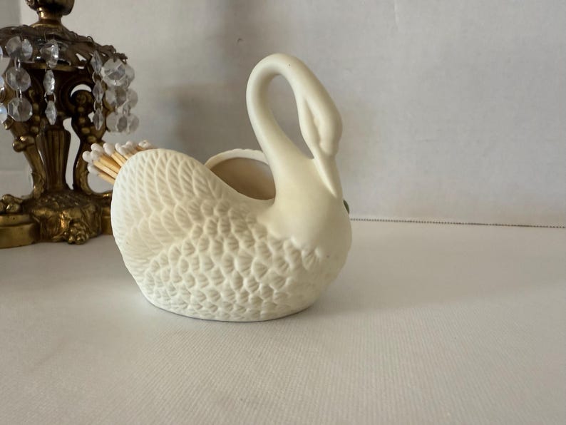 May include: Cream-colored ceramic swan figurine with textured feather detailing. The swan has a curved neck and a small opening on its back. A decorative brass and crystal candle holder is partially visible in the background.