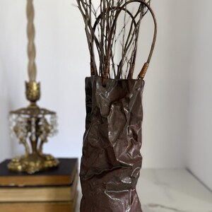May include: A tall, dark brown ceramic vase with a textured, crumpled paper-like surface. The vase features two thin, arched handles attached to the top rim and contains a cluster of thin, bare, brown twigs extending upward from the opening.