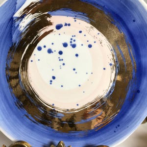 May include: A white ceramic plate with a blue and gold design. The plate has a circular pattern with a pink center and blue dots. The plate is on a gold metal stand.