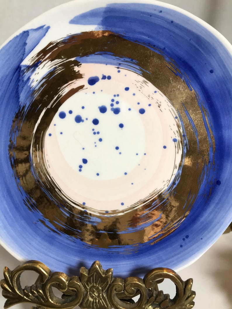 May include: A white ceramic plate with a blue and gold design. The plate has a circular pattern with a pink center and blue dots. The plate is on a gold metal stand.