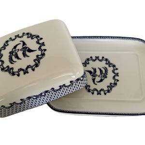Op de afbeelding: Crèmekleurige keramische botervloot met bijpassend deksel. Beide stukken hebben een donkerblauwe decoratieve rand en een centraal ontwerp van gestileerde vogels in een cirkelvormige krans. Het deksel staat iets open.