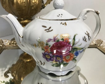 May include: White porcelain teapot with a floral design in shades of pink, yellow, orange, and blue. The teapot has a gold trim and a lid with a knob.