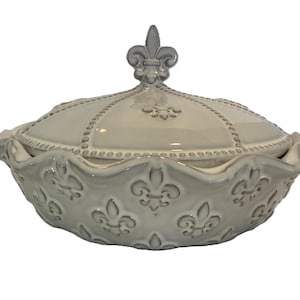 Puede incluir: Una fuente de cerámica gris claro con tapa. La fuente presenta un borde festoneado y está adornada con diseños de flor de lis. La tapa está rematada con un remate de flor de lis y tiene un borde con cuentas. Una pieza decorativa para la cocina o el comedor.