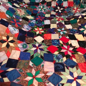 May include: A colorful patchwork quilt with a starburst pattern. The quilt is made of various fabrics in shades of red, blue, green, and brown. The quilt is stitched together with red thread.
