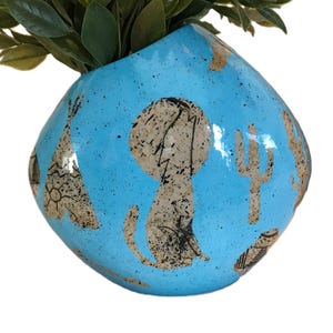 Op de afbeelding: Een turquoise keramische vaas met een gespikkelde afwerking, met bruine illustraties van een tipi, cactus en andere woestijnmotieven. De vaas is gevuld met groene, bladrijke stelen.