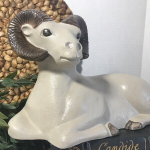 Op de afbeelding: Een wit keramiek beeld van een ram met bruine hoorns. De ram ligt met zijn hoofd naar de zijkant gedraaid. Het beeld staat op een zwart boek met het woord "Candide" erop.