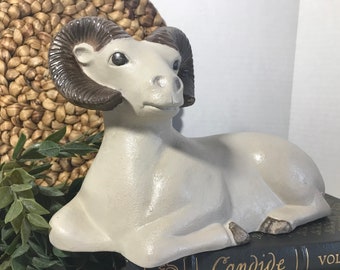May include: A white ceramic sculpture of a ram with brown horns. The ram is lying down with its head turned to the side. The sculpture is on a black book with the word "Candide" visible.