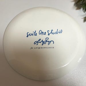 May include: White ceramic plate with blue text that reads "Suite One Studio" and a blue signature that reads "Emily Sory" followed by "for ANTHROPOLOGIE".