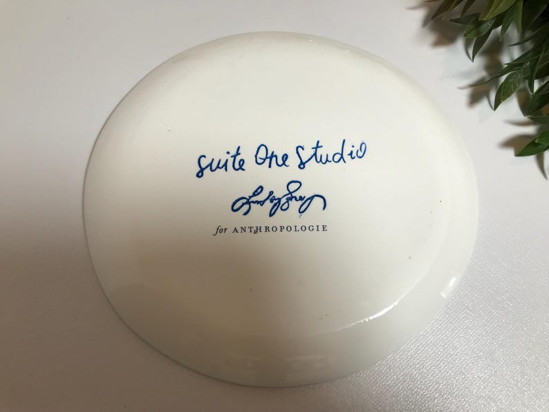 May include: White ceramic plate with blue text that reads "Suite One Studio" and a blue signature that reads "Emily Sory" followed by "for ANTHROPOLOGIE".