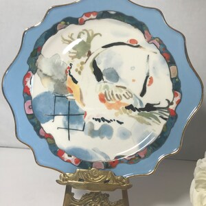 May include: A small, scalloped, blue and white porcelain plate with a gold rim. The plate features a watercolour painting of a bird in flight with a blue and white background. The plate is displayed on a gold metal easel.