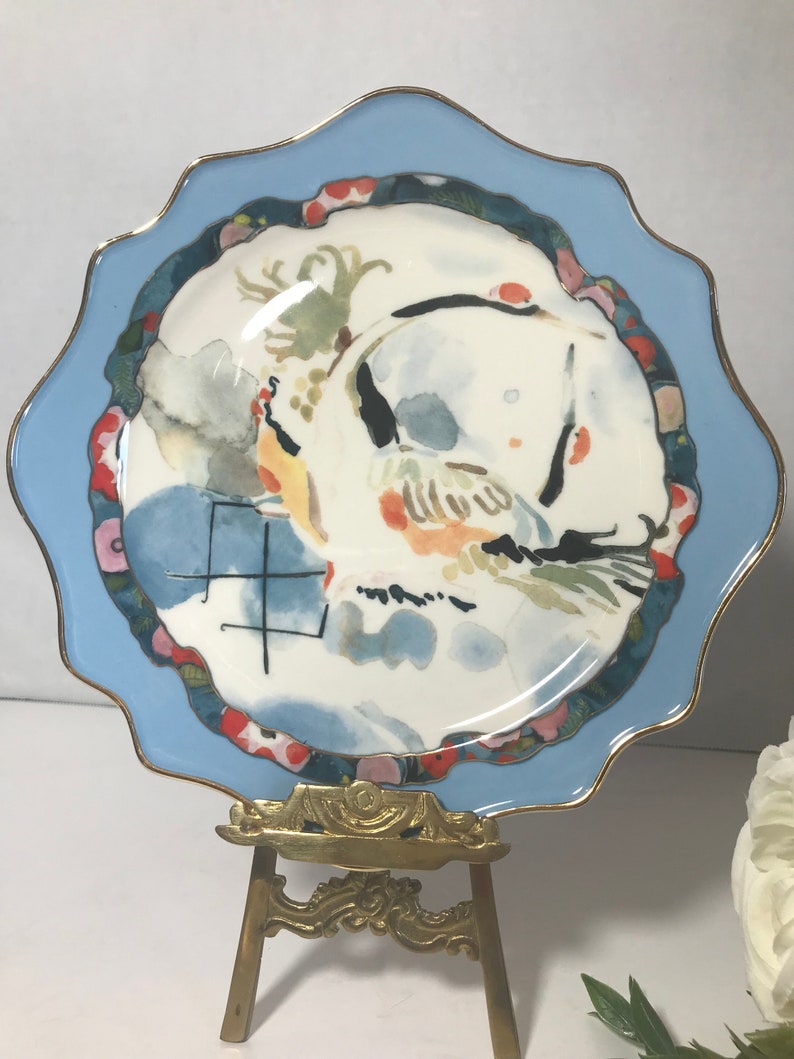 May include: A small, scalloped, blue and white porcelain plate with a gold rim. The plate features a watercolour painting of a bird in flight with a blue and white background. The plate is displayed on a gold metal easel.