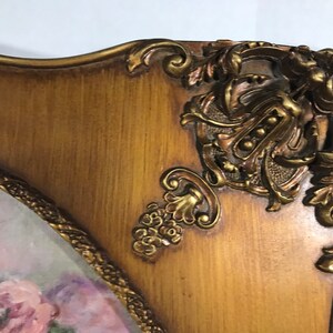May include: A gold-toned ornate picture frame with a floral painting inside. The frame has intricate details and a wood-like finish.
