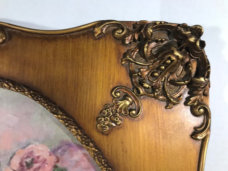 May include: A gold-toned ornate picture frame with a floral painting inside. The frame has intricate details and a wood-like finish.