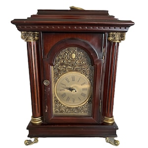 May include: A dark wood mantel clock with a gold-colored clock face and decorative accents. The clock has Roman numerals and ornate detailing on the front and sides. The clock is rectangular with a decorative top and base.