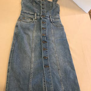 Peut inclure: Une robe en jean bleu avec un devant boutonn&eacute; et des bretelles fines. La robe a un corsage ajust&eacute; et une jupe l&eacute;g&egrave;rement &eacute;vas&eacute;e.