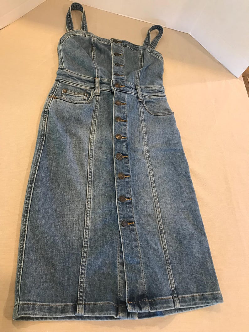 Peut inclure: Une robe en jean bleu avec un devant boutonn&eacute; et des bretelles fines. La robe a un corsage ajust&eacute; et une jupe l&eacute;g&egrave;rement &eacute;vas&eacute;e.